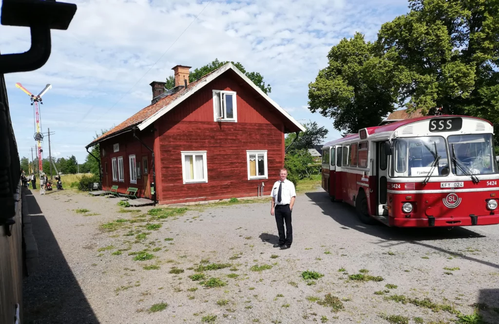 Bärby station på Lennakatten, ett tåg gör uppehåll, en röd-vit buss och en busschaufför står på stationsplanen.