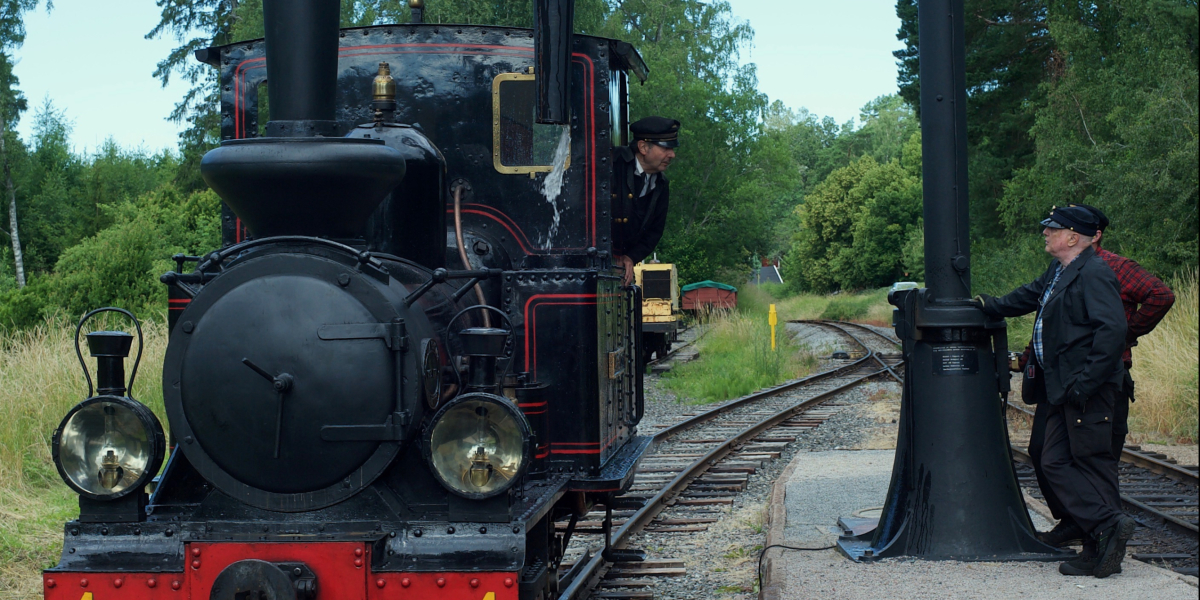 Lennakattens lilla ånglok tankar vatten medan järnvägspersonal ser på.