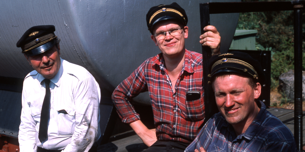 Tre järnvägsarbetare i skjorta och skärmmössa sitter och har trevligt på en tankvagn.