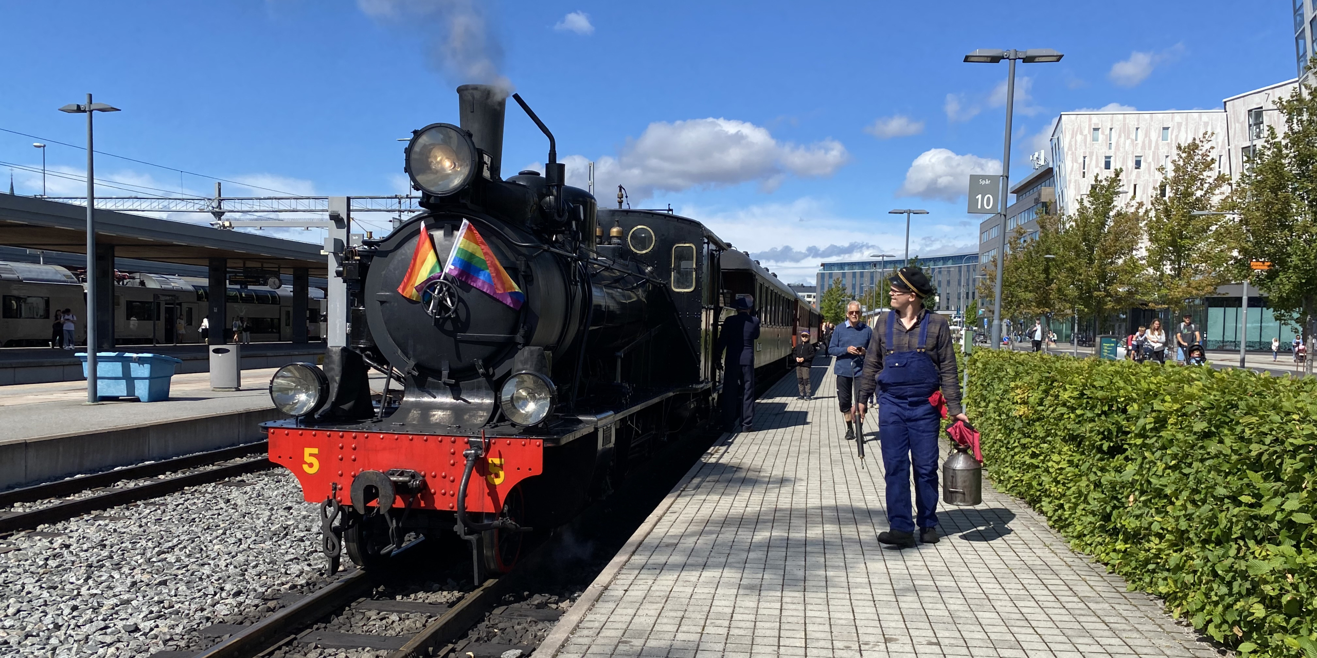 Ånglok som pryds av två prideflaggor i fronten. Lokföraren står bredvid med en oljekanna.