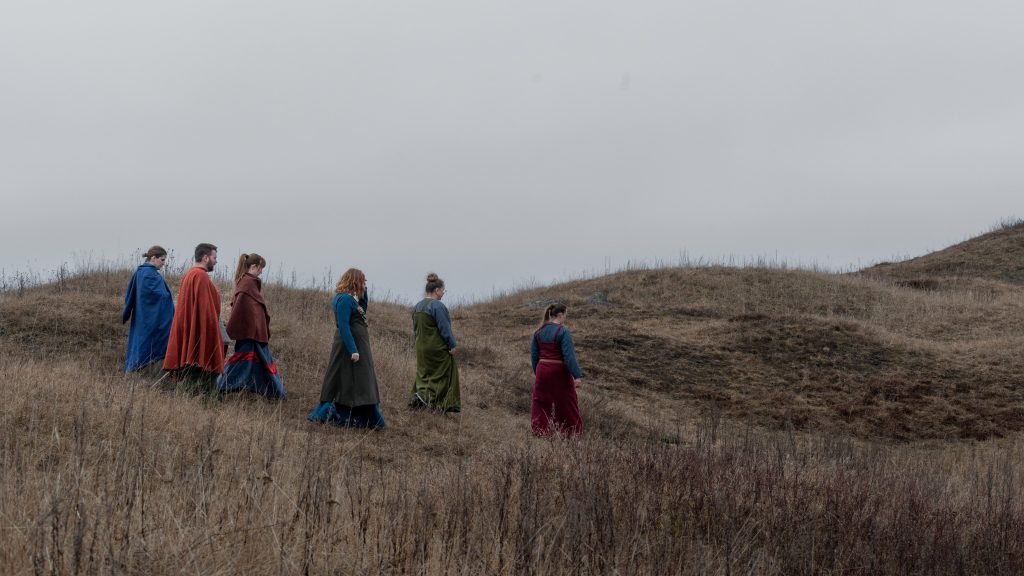 Vikingar går i grupp längs kullar. Vädret är mulet.